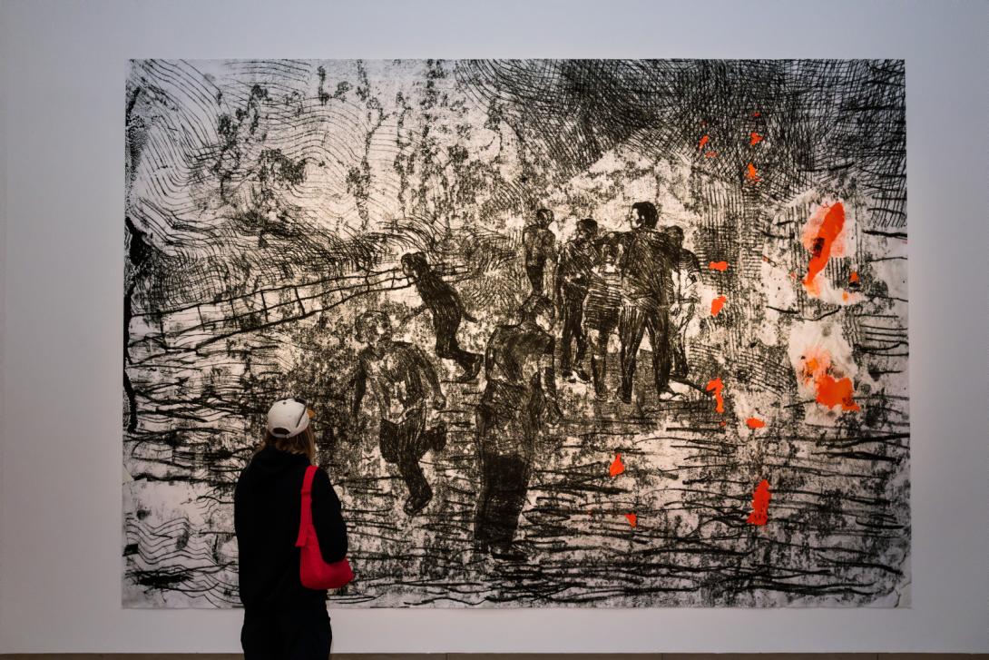 A very large print in black, white and orange tones installed on a white gallery wall, with a visitor looking on