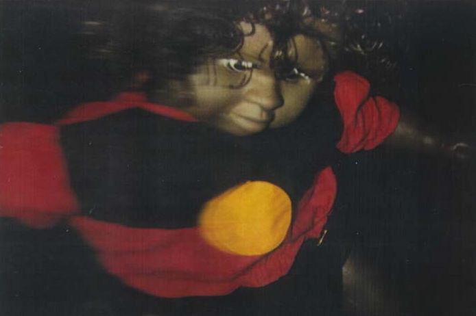A detail view of a photograph depicting a baby doll with a dark skin tone, wearing a doll-sized sweater emblazoned with the Australian Aboriginal flag