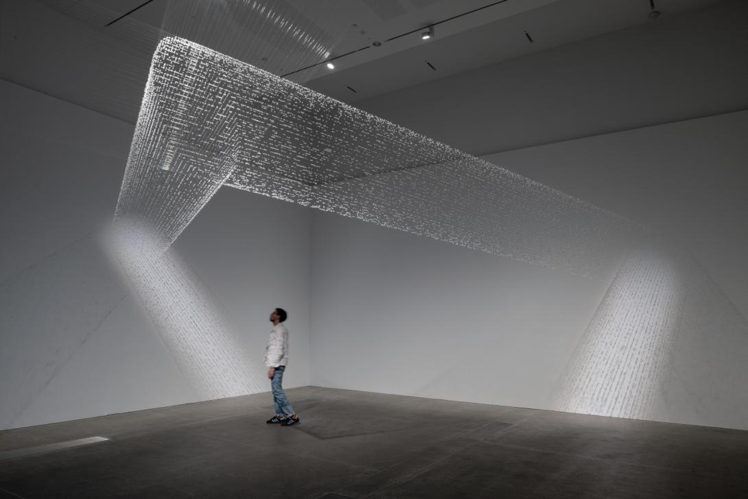 A view of a delicate spiderweb-like installation in a dimmed gallery space, with a visitor standing underneath the work.