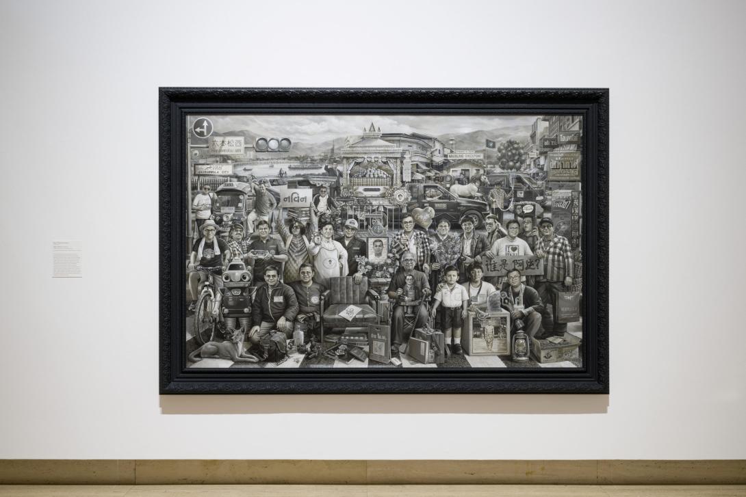 A large work rendered in black and grey tones installed on a white wall in a gallery space.