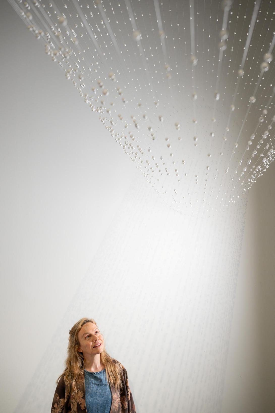 A photograph in a bright gallery space of the artist Sandra Selig standing underneath her installation mid-air 2003