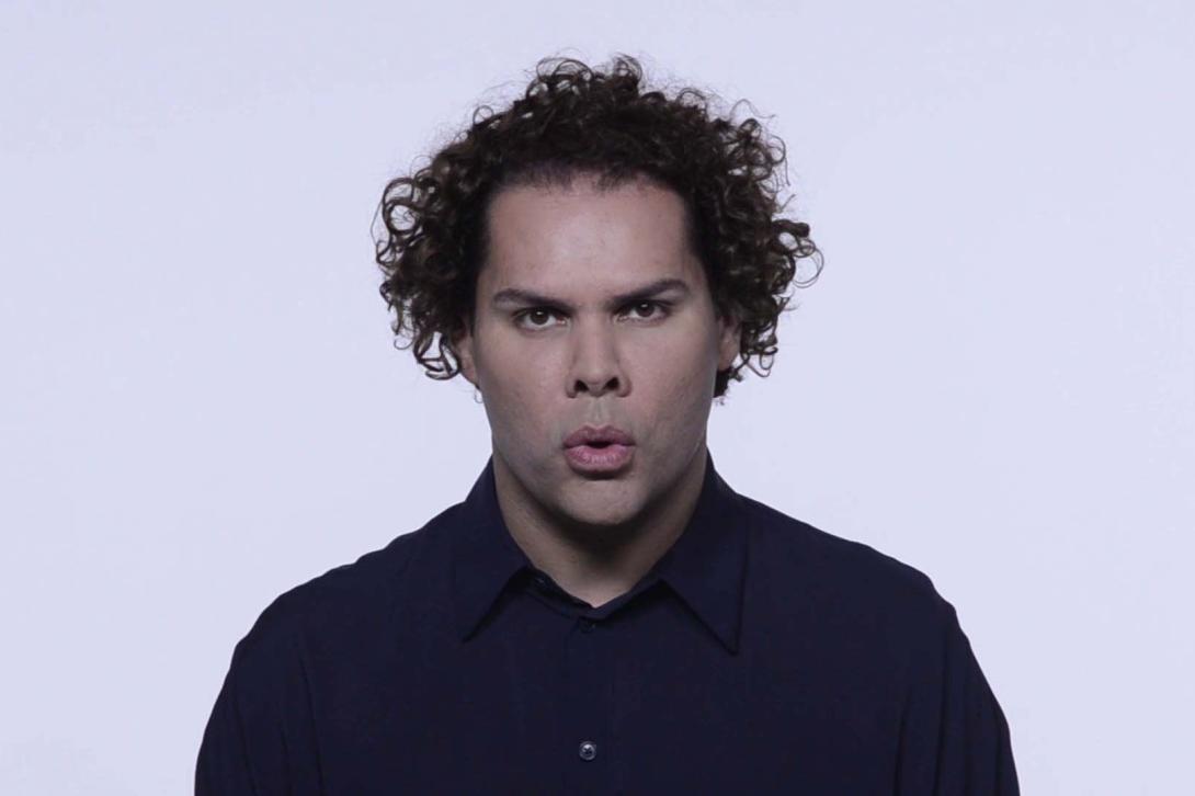 A still drawn from a video work, in which we see the artist Dr Christian Thompson – an artist of Bidjara/Irish/Chinese–Australian heritage – standing in front of a bright background. He appears to be in the middle of singing. He has short, curly hair and wears a dark blue button-up shirt.