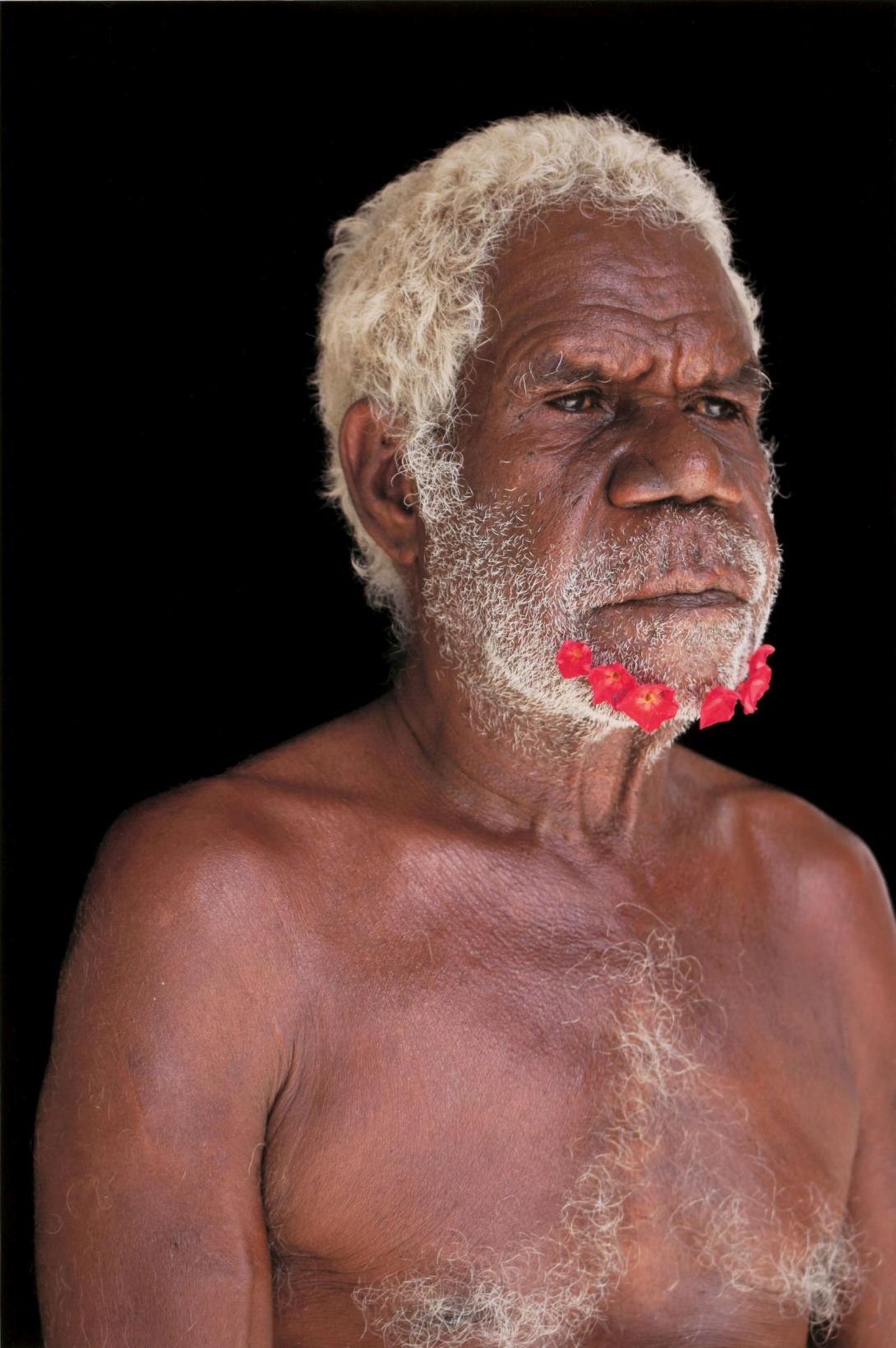 A portrait-style photograph of an older Indigenous Australian man, with white hair and with red flowers adorning his cropped white beard. 
