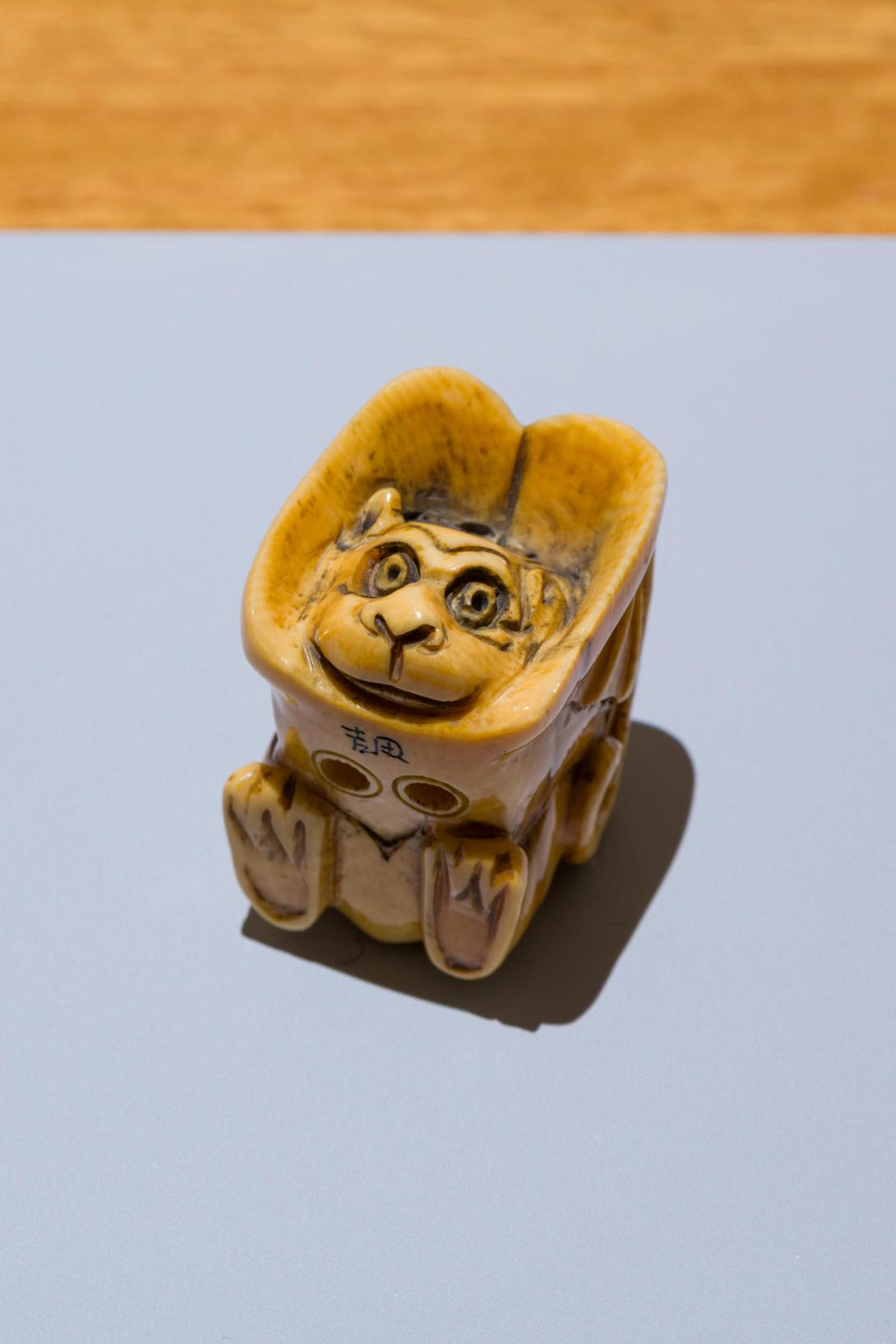 A photograph of a tiny bat, carved from ivory, with its wings wrapped around its body. The bat has a cheeky smile and bright eyes. Its little feet are visible, carved on the figure's underside.