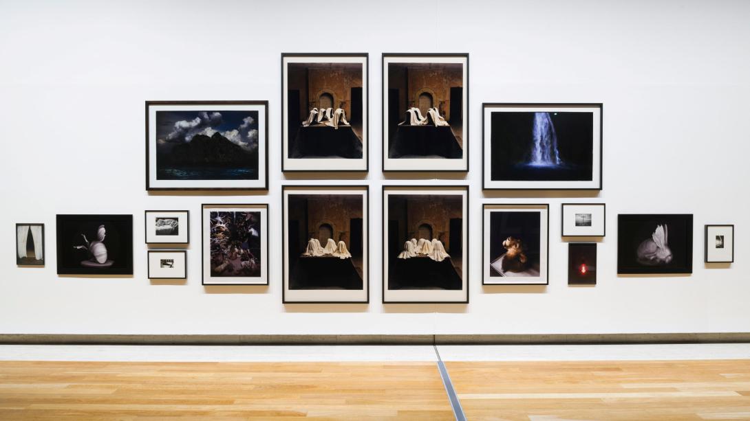 This photograph captures a view of a salon-style installation of photographs, with very small works hung at the far edges and the largest works hung in the middle. Multiple artists are represented; however, each photograph included has dramatic, Baroque elements in its composition.