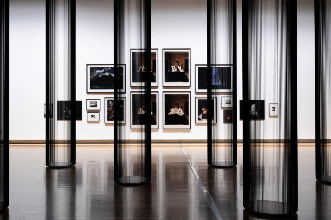 This photograph captures a view of a salon-style installation of photographs, with very small works hung at the far edges and the largest works hung in the middle. In the foreground, columns made of tape from inside VHS cassettes appear both monumental and transparent. Multiple artists are represented; however, each photograph included has dramatic, Baroque elements in its composition.