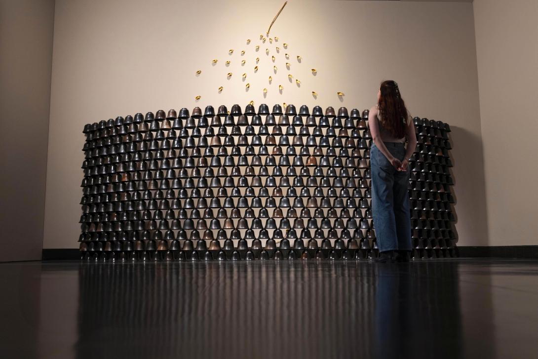 An installation view of a sculptural work appearing as a curved wall of ceramic bells; a person stands before the work, viewing it