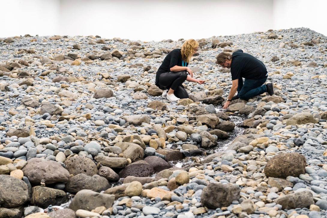 In this photograph, a person crouches on a rocky surface and extends a hand into a thin stream of running water that winds between grey and tan stones in a white-walled gallery.