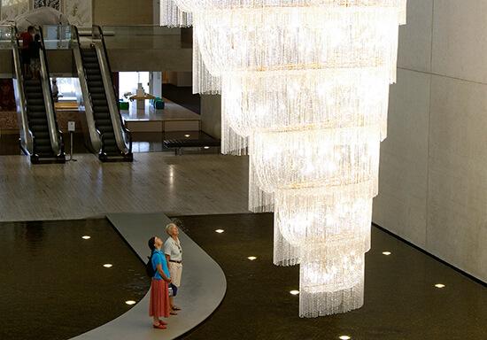 Ai Weiwei, China b.1957 / Boomerang 2006 / Glass lustres, plated steel, electric cables, LED lamps / 700 x 860 x 290cm / Gift of the artist through the Queensland Art Gallery Foundation 2007 / Collection: Queensland Art Gallery | Gallery of Modern Art / © Ai Weiwei / Photographs: Natasha Harth & Joe Ruuckli © QAGOMA Two people in the Gallery's water mall look up at a huge, elaborate chandelier.
