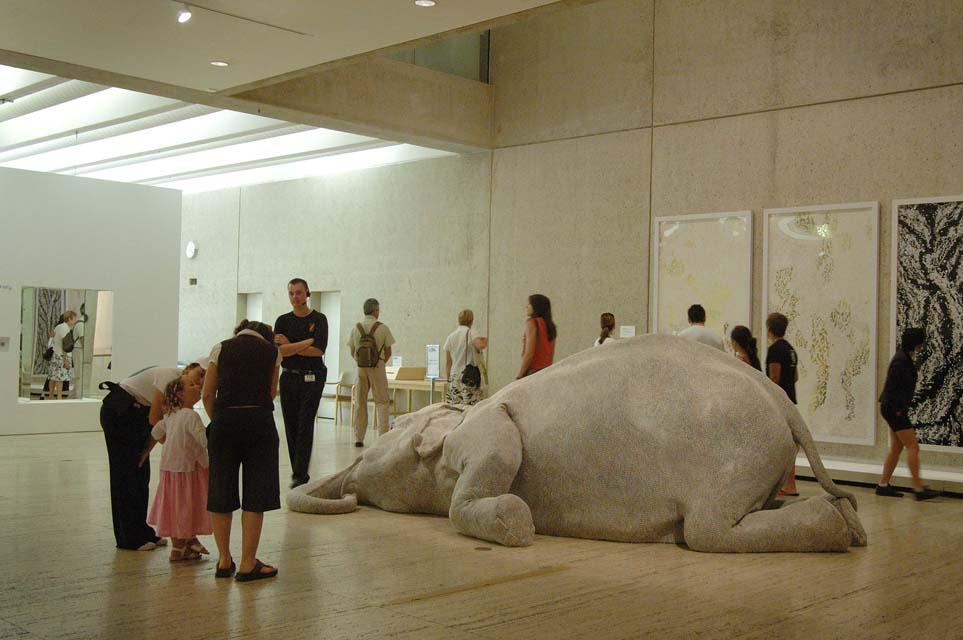 Bharti Kher’s sculpture The skin speaks a language not its own 2006, installed at QAG for APT5, 2006 An installation view of a life-size and very realistic sculpture of an elephant, slumped to the floor as if dead, in a gallery space surrounded by onlookers.