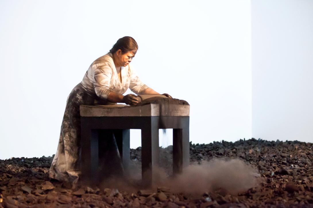 In a gallery space, a woman crushes and grinds hundreds of kilograms of charcoal briquettes into powder and dust