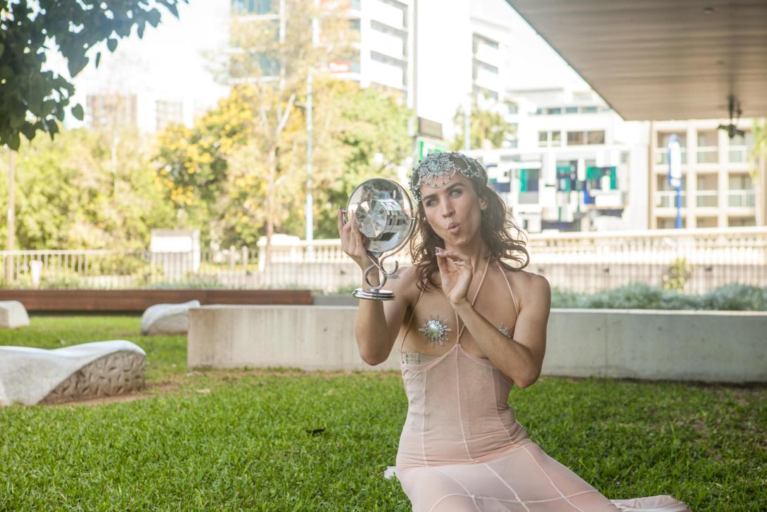 A dancer costumed in a sparkly, form-fitting cream dress and tiara looks into a round mirror; she is seated outside of GOMA on the grass.