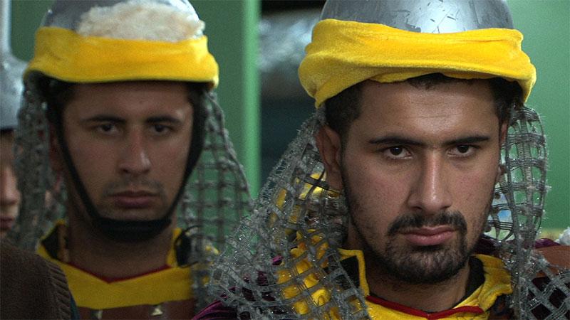 A still photo of two men wearing costumes comprising a hat with a yellow band and leather armour.