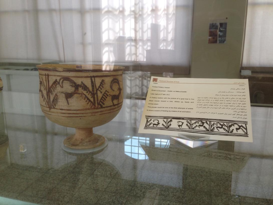 An earthenware goblet on display with a descriptive plaque behind it, with the reflection of white curtains on the cabinet glass.