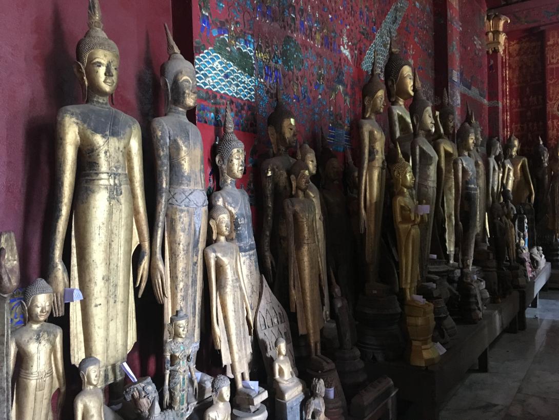 Gold-toned Buddhist statues stand in a grouping against a red wall.