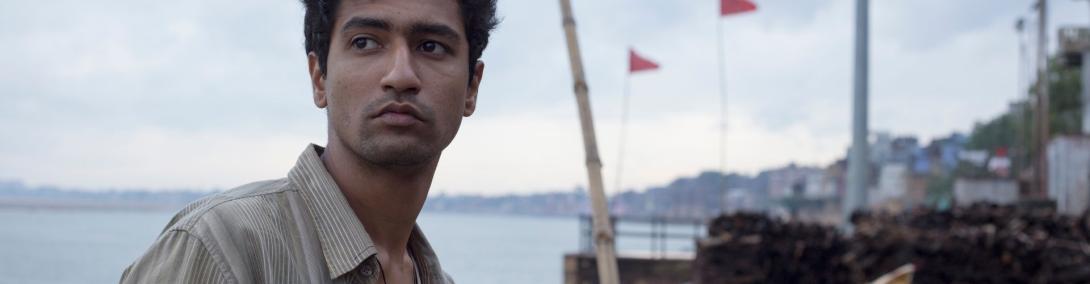 A still image of a man looking past the camera; behind him is a ship on the ocean.