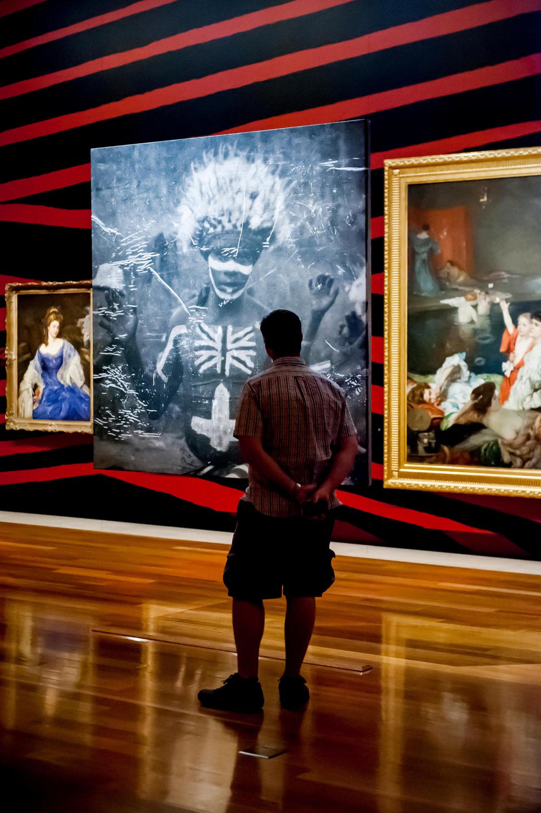 An installation view of Brook Andrew's Intervening Time, featuring a work from Andrew's 'TIME' series at centre, with works from QAGOMA's Australian Collection (including Rupert Bunny's Bathers) at either side. The wall is painted in black and red stripes.