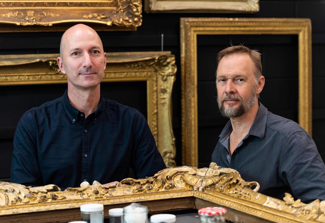 Two men stand in the QAG Framing Workshop, with gold-gilded frames on a black wall behind them and a large gold-gilded frame on a table before them.