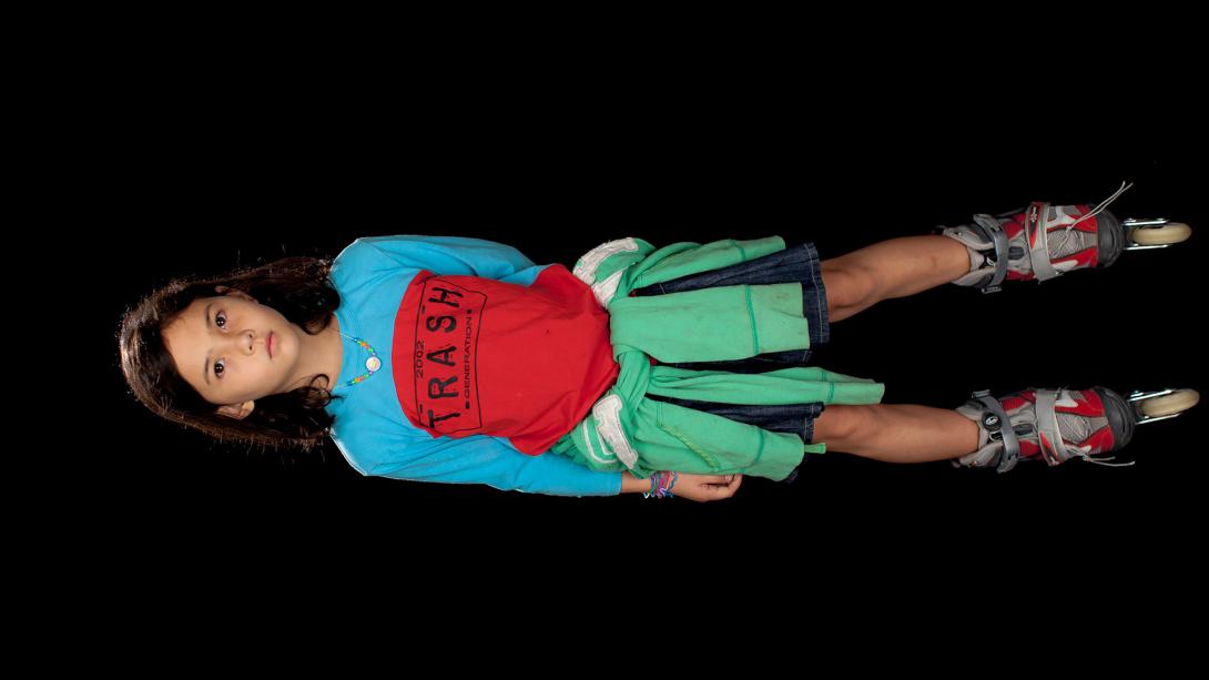 A photograph on its side of a young girl wearing a red-and-blue shirt, green sweater tied around her waist, and red rollerblades. She looks directly at the camera.