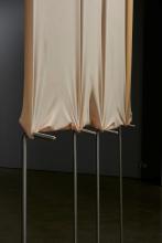 An installation view of a sculptural work that appears as a beige latex curtain suspended on a steel frame in a concrete room.