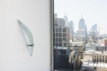 A detail view of a work made of shattered glass, installed to the left of a window, through which one can see blue sky and a city view.