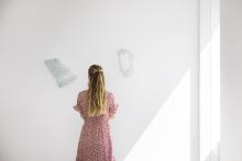 A detail view of works made of shattered glass installed on a white wall, with a viewer in a red-and-white dress looking at them closely.
