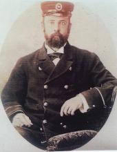 A sepia photograph of a man in a late-1800s naval uniform, with a short and tidy beard, and a hat.