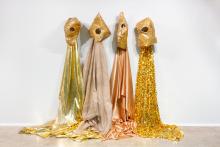 An installation view of four costume-like sculptures made of various shades of gold fabric, which are suspended from a white wall and trail down onto the floor.