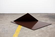 An abstract artwork made of steel appears as a bronze-coloured sheet of metal with one corned turned up; it is installed on a concrete floor.