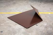 An abstract artwork made of steel appears as a bronze-coloured sheet of metal with one corned turned up; it is installed on a concrete floor.