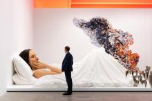 An installation view of a realistic sculpture of a giant woman lying in bed, with a visitor looking on.