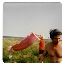 A square colour photograph of a man standing in a field, with a person behind him unfurling a red blanket.