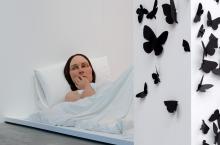 An installation view of a realistic sculpture of a giant woman lying in bed.