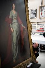 An oil painting of a woman in a blue dress and red robe, with an ornate gold frame, supported by conservators about to remove its frame.