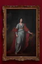 An oil painting of a woman in a blue dress and red robe, with an ornate gold frame, hung on a dark red wall.