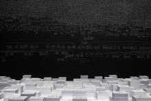 An installation view of a dark gallery space, where the artwork is created in white chalk over the black walls; the centre of the space includes an elevated platform covered in stacks of paper