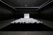 An installation view of a dark gallery space, where the artwork is created in white chalk over the black walls; the centre of the space includes an elevated platform covered in stacks of paper