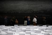An installation view of a dark gallery space, where the artwork is created in white chalk over the black walls; the centre of the space includes an elevated platform covered in stacks of paper