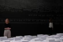 An installation view of a dark gallery space, where the artwork is created in white chalk over the black walls; the centre of the space includes an elevated platform covered in stacks of paper