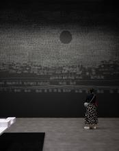 An installation view of a dark gallery space, where the artwork is created in white chalk over the black walls; the centre of the space includes an elevated platform covered in stacks of paper