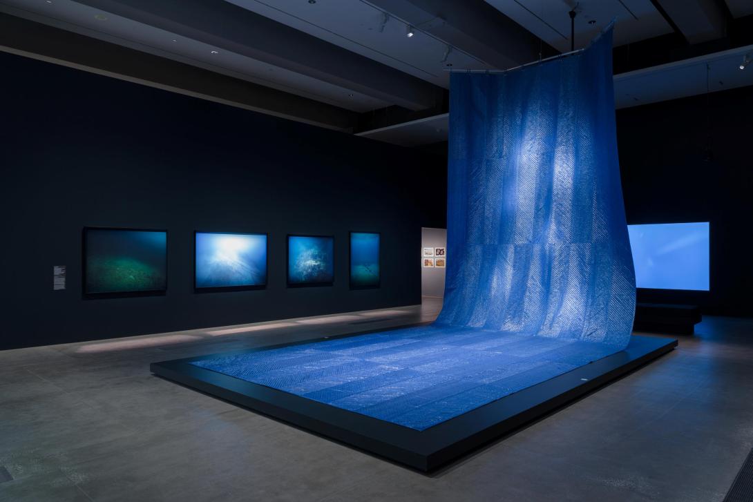 An installation view of a dim gallery space that features a large, blue, wave-like work, with photographs behind on the dark walls.