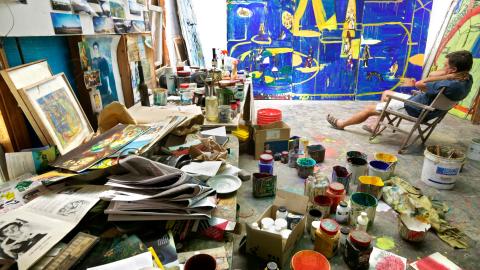 A photograph of an artist sitting in his studio, surrounded by paint cans, with a huge blue-painted canvas against the back wall.