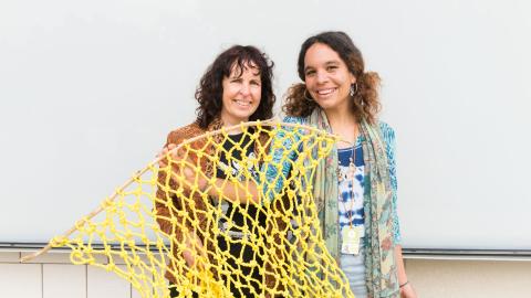 Two women stand in front of GOMA's external wall holding a yellow fishing net and smiling.