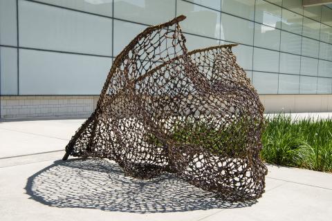 An installation view of a work of public art photographed on a sunny day; it takes the form of a large fishing net, cast in bronze, and it is set in front of GOMA, near the grass. It casts a net-like shadow on the concrete.