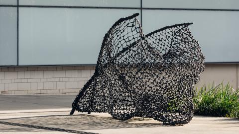 A daylight view of a sculpture, taking the form of a fishnet cast in bronze, installed outside of GOMA