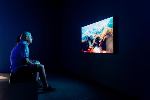 A photograph of a dark gallery space in which a seated person watches a screen, which displays an underwater scene.