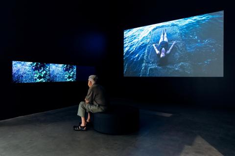 An installation view of a three-channel and a single-channel video work playing in a dark gallery space while a seated visitor watches.
