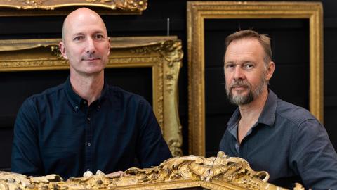Two men stand in the QAG Framing Workshop, with gold-gilded frames on a black wall behind them and a large gold-gilded frame on a table before them.