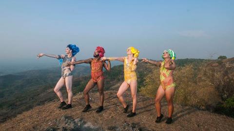 In this photograph, four drag queens stand on a high hill overlooking a valley; they wear blue, red, yellow and green respectively.