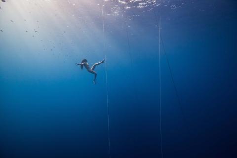A still photograph taken underwater of a feminine-appearing person swimming nude in deep, clear blue water. The photo is taken from a distance underwater.